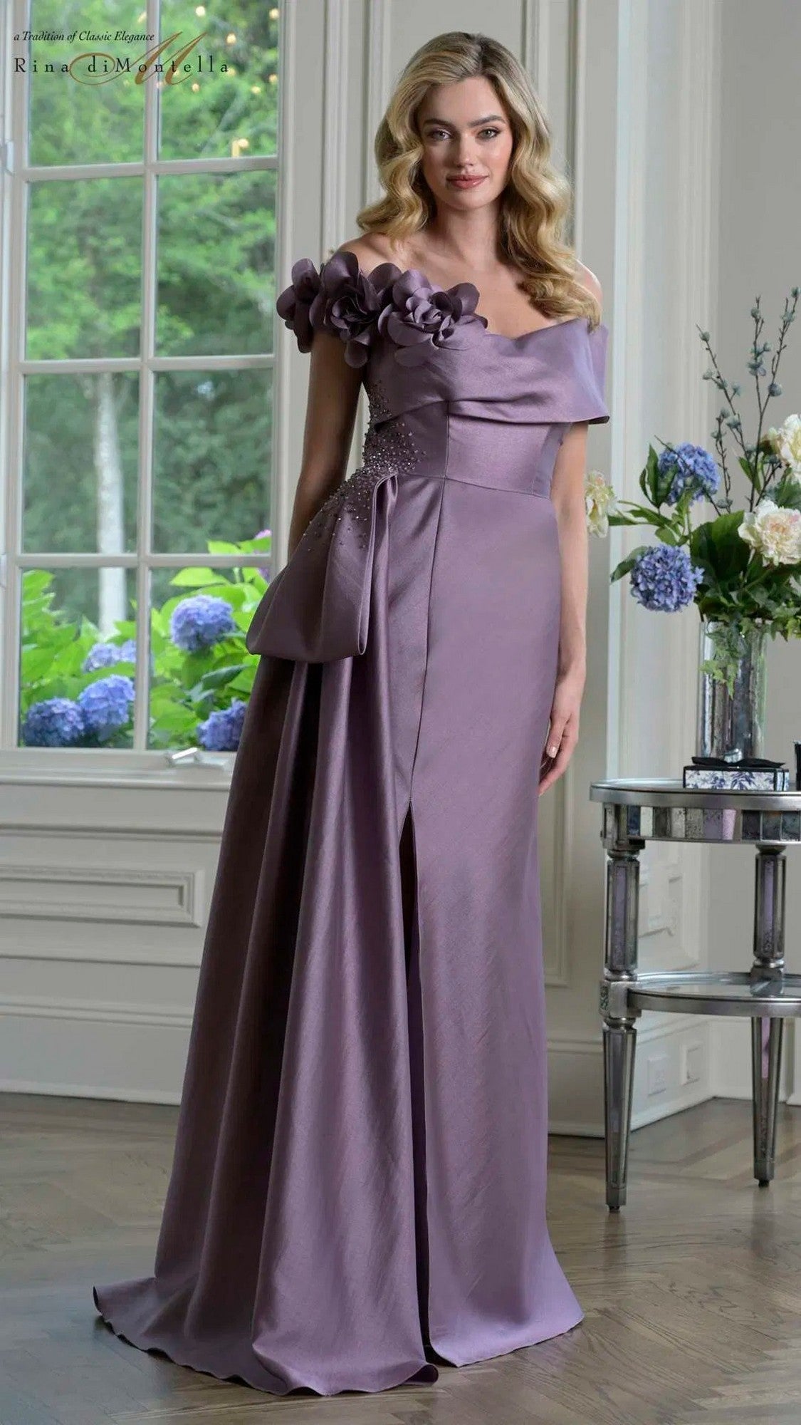 Indoors, a woman wears the Rina Di Montella RD3407 Long Formal Dress in Victorian Lilac, featuring floral shoulder details and a large bow, standing beside a table with vases of flowers.